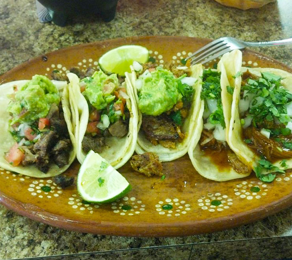 Five tacos with various fillings, topped with guacamole and cilantro, served on a decorative plate with lime wedges.