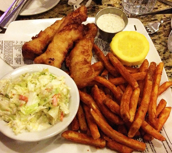 Plate of fried fish, French fries, coleslaw, a lemon wedge, and a cup of tartar sauce on a table.