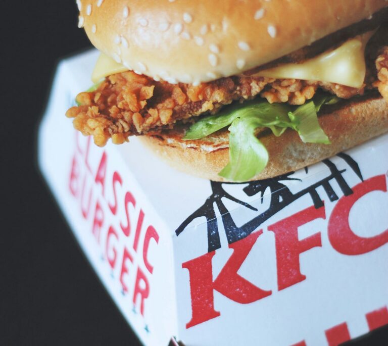 Close-up of a KFC chicken sandwich with lettuce and cheese on a sesame bun, resting on a branded box.