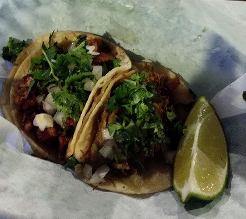 Two street-style tacos topped with cilantro and onions, served with lime wedges on parchment paper.