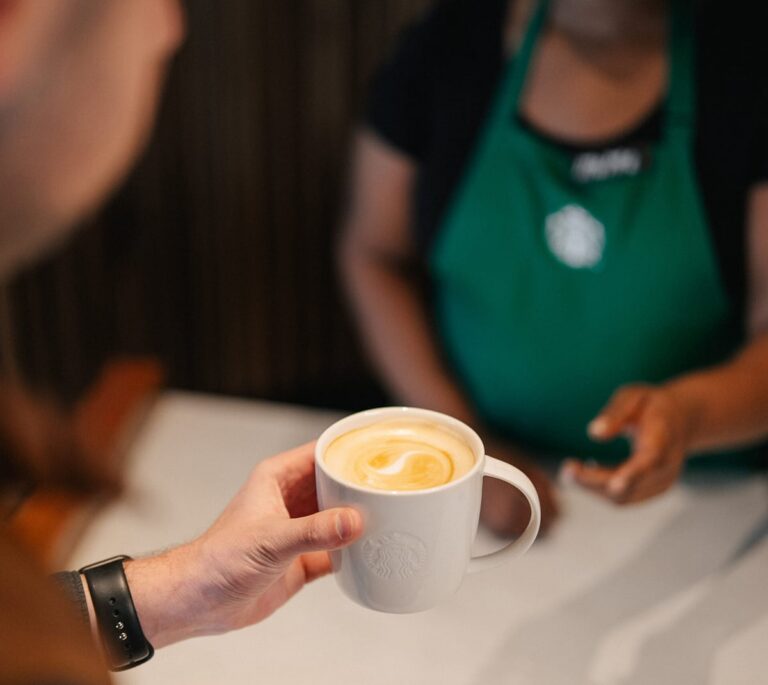 A person holds a cup of coffee at a counter, served by a barista wearing a green apron.