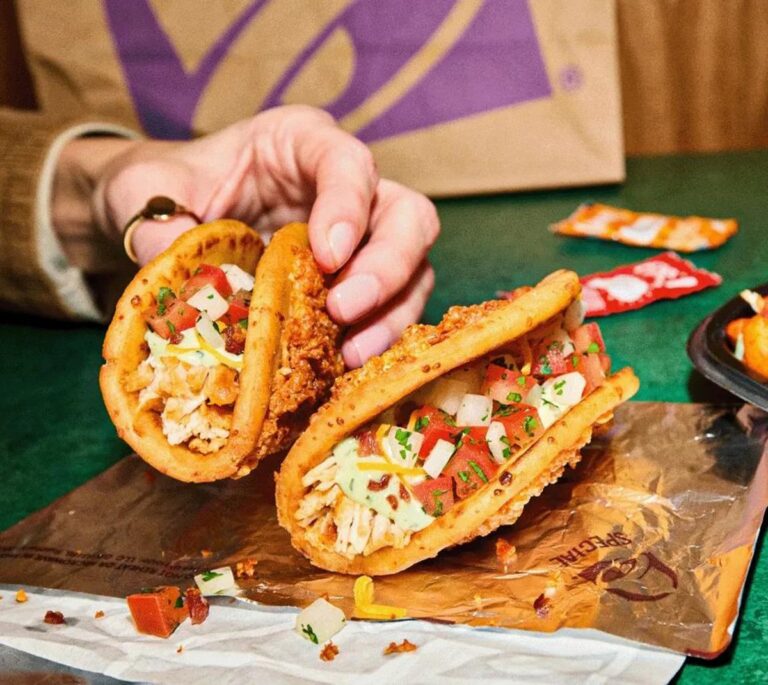 A hand holds a Taco Bell taco with a fried chicken shell, filled with lettuce, tomato, and sauce.