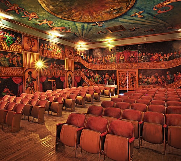 Theater with red seats, ornate murals on walls and ceiling, and dramatic lighting, no audience present.