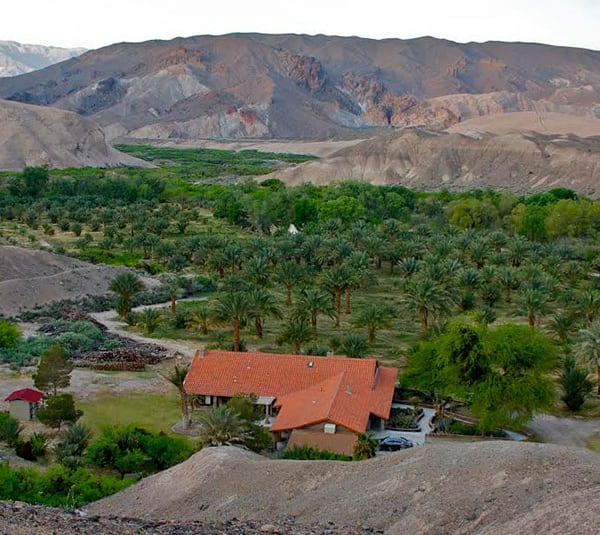A house with a red roof sits among palm trees in a green oasis surrounded by arid, rocky hills.
