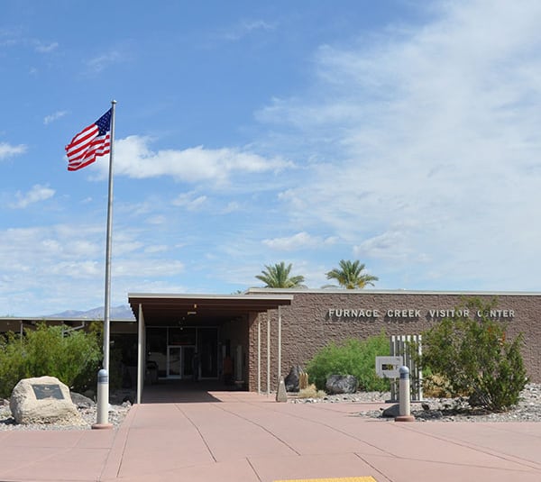 An American flag flies outside the Furnace Creek Visitor Center under a blue sky with scattered clouds.