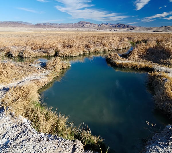 A small clear pond in a grassy, dry landscape with distant mountains under a blue sky with wispy clouds.
