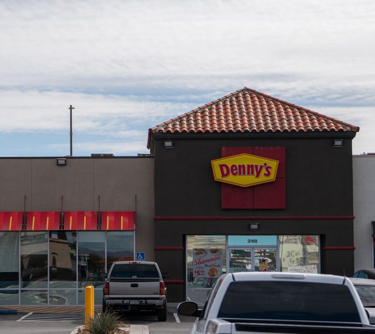 Dennys restaurant exterior with a tiled roof and parked cars in front under a cloudy sky.