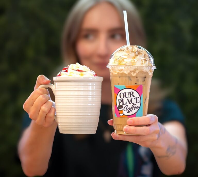 A person holds a mug of whipped cream-topped coffee and an iced coffee labeled “Our Place Coffee.”.