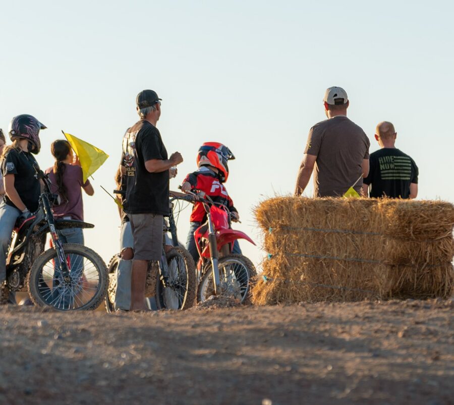 People stand near dirt bikers and hay bales on a dirt track, with yellow flags and a clear sky in the background.