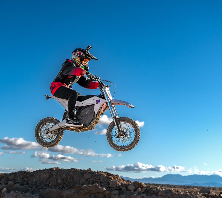 Person riding a dirt bike mid-air over a dirt track with a clear blue sky and mountains in the background.