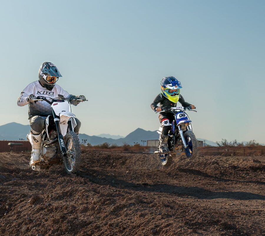 An adult and a child ride dirt bikes on a dirt track with mountains in the background.