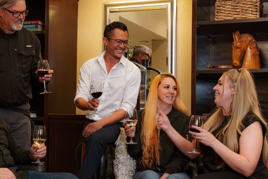 Five adults laughing and talking while holding wine glasses in a cozy room with shelves and warm lighting.