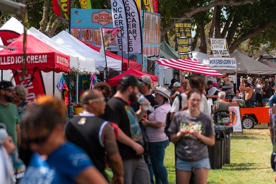 Crowd walking among colorful food and vendor tents at an outdoor fair on a sunny day.