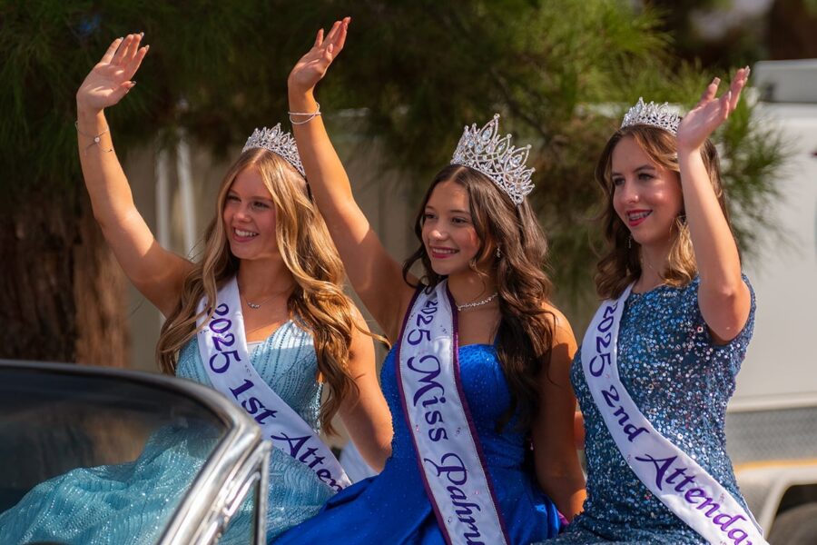 Three young women in crowns and sashes wave while riding in a parade, dressed in sparkly blue gowns.