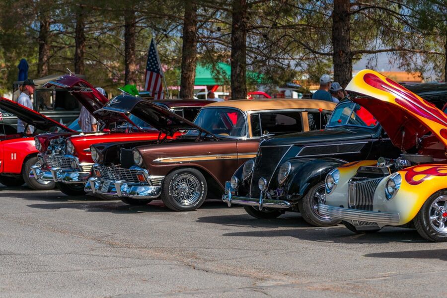 Classic cars with open hoods on display at an outdoor car show, trees and people in the background.