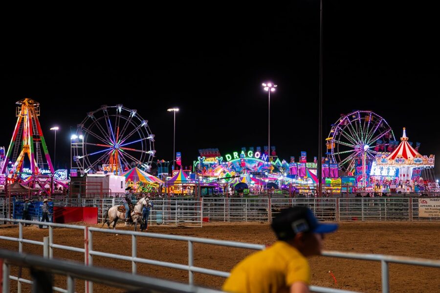 Nighttime rodeo scene with two horses, Ferris wheels, and colorful carnival rides in the background.