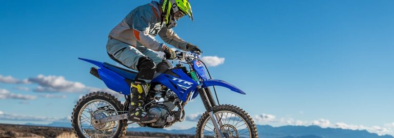Person wearing gear and helmet riding a blue dirt bike over rough terrain under a clear blue sky.