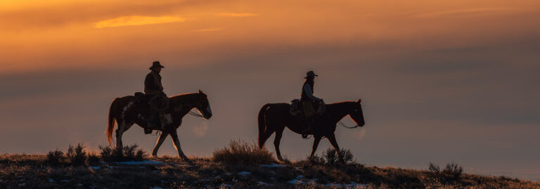 Two people on horseback ride along a ridge at sunset, silhouetted against an orange sky.