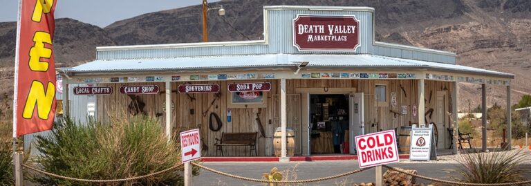 Death Valley Marketplace storefront with signs for local crafts, souvenirs, cold drinks, and scenic mountain backdrop.