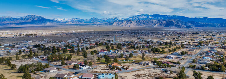Aerial view of a small town with houses, trees, and mountains in the background under a partly cloudy sky.
