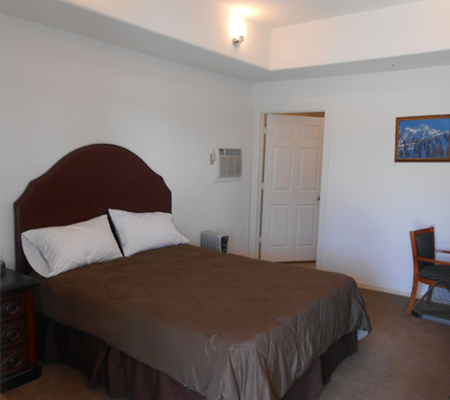 A neatly made bed with brown bedding in a simple, clean bedroom with white walls and minimal decor.