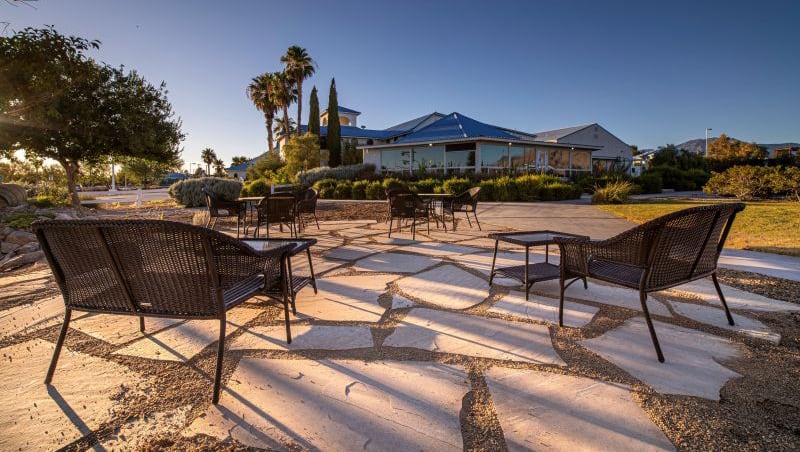 Outdoor patio with black metal chairs and tables on stone pavement, surrounded by trees and greenery.