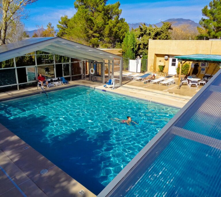 A person swims in a clear, covered outdoor pool with lounge chairs and trees around.