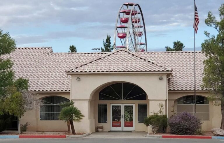 A ferris wheel rises behind a beige building with a tiled roof and an American flag on a pole.