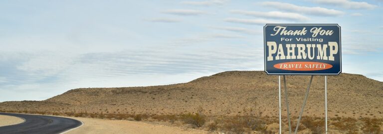 Roadside sign reads Thank You For Visiting Pahrump, Travel Safely in a desert landscape with hills.