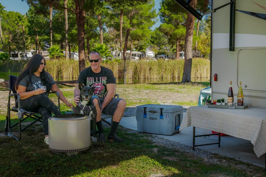 A man and woman relax with a dog by a camper, sitting near a fire pit and a table with drinks outdoors.