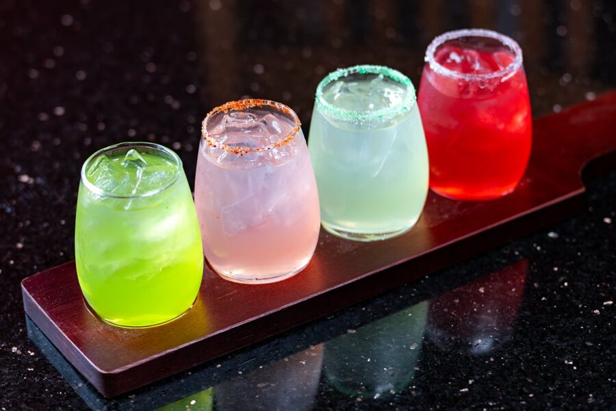 Four colorful cocktails in rounded glasses on a wooden tray against a dark, reflective surface.