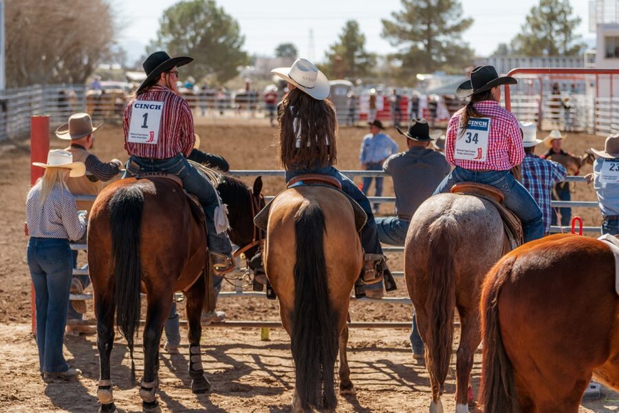 Three rodeo participants on horseback, wearing numbered shirts and cowboy hats, wait by an arena fence.