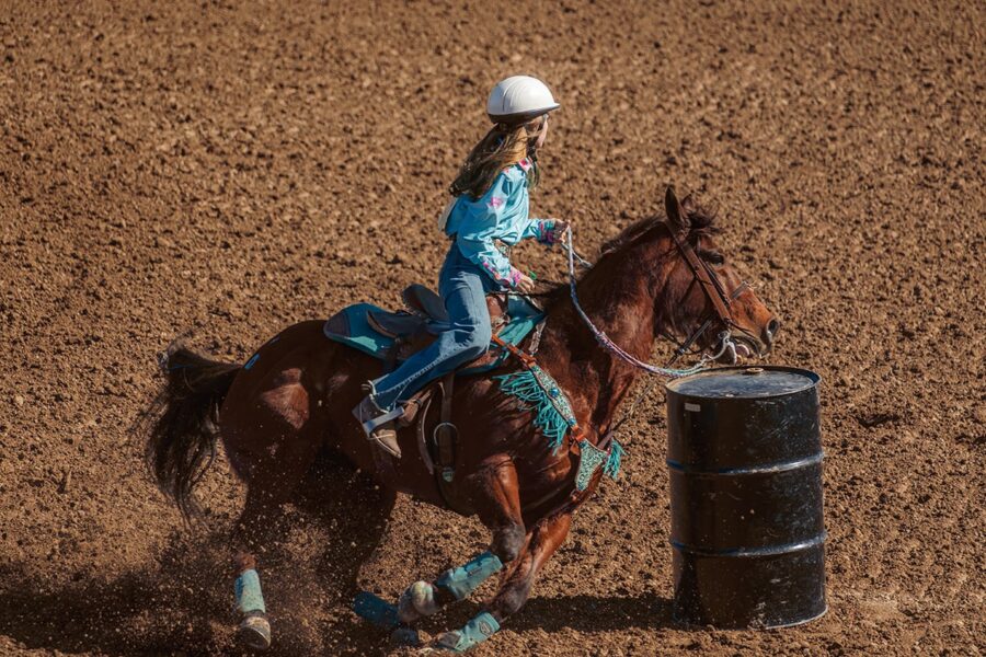 A girl in a helmet rides a horse around a barrel during a rodeo barrel-racing event on dirt.