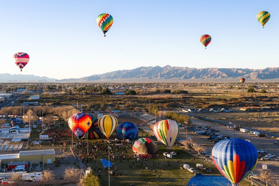 Colorful hot air balloons rise over a festival field with mountains in the background under a clear sky.