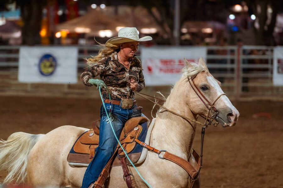 A woman in a cowboy hat rides a light-colored horse at a rodeo event in an arena.