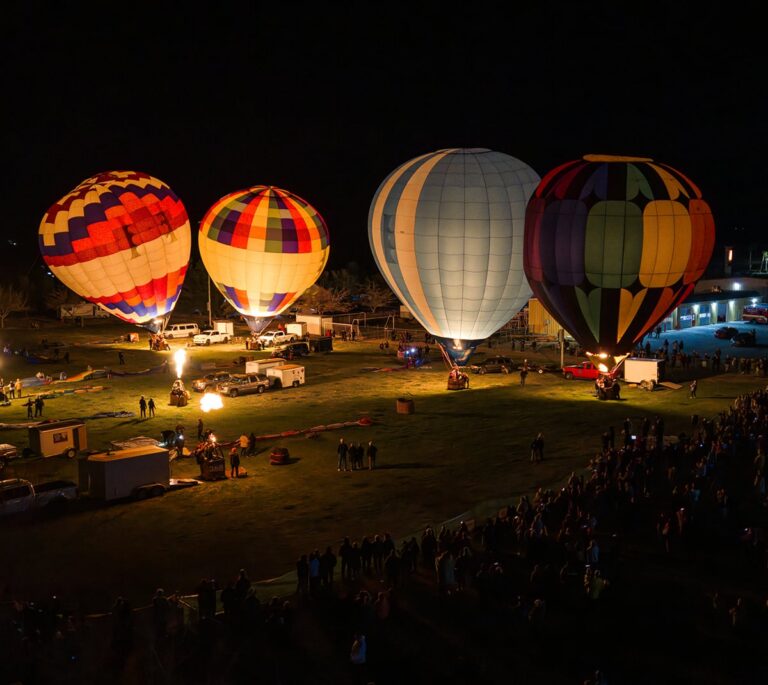 Four colorful hot air balloons glowing at night, surrounded by a crowd on a grassy field.