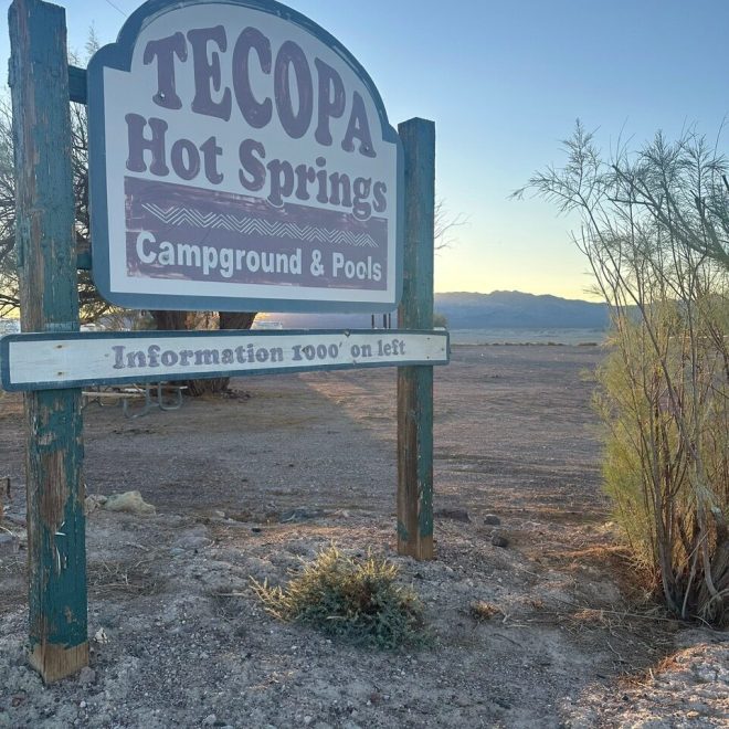 Sign for Tecopa Hot Springs Campground & Pools; desert landscape and mountains in the background.